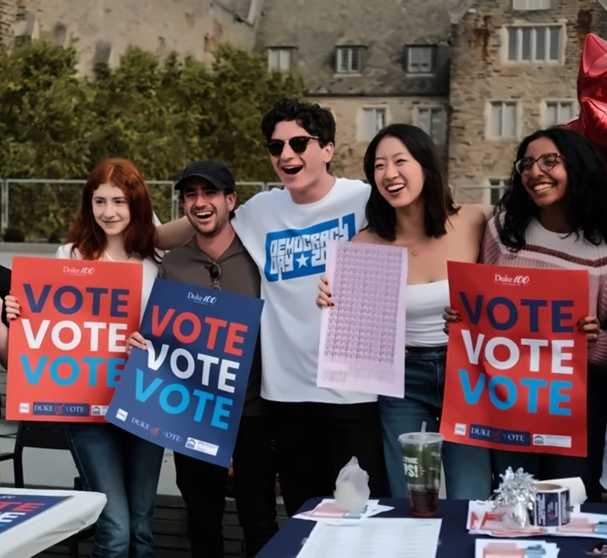 Students laughing together at a voter registration table, holding Vote signs